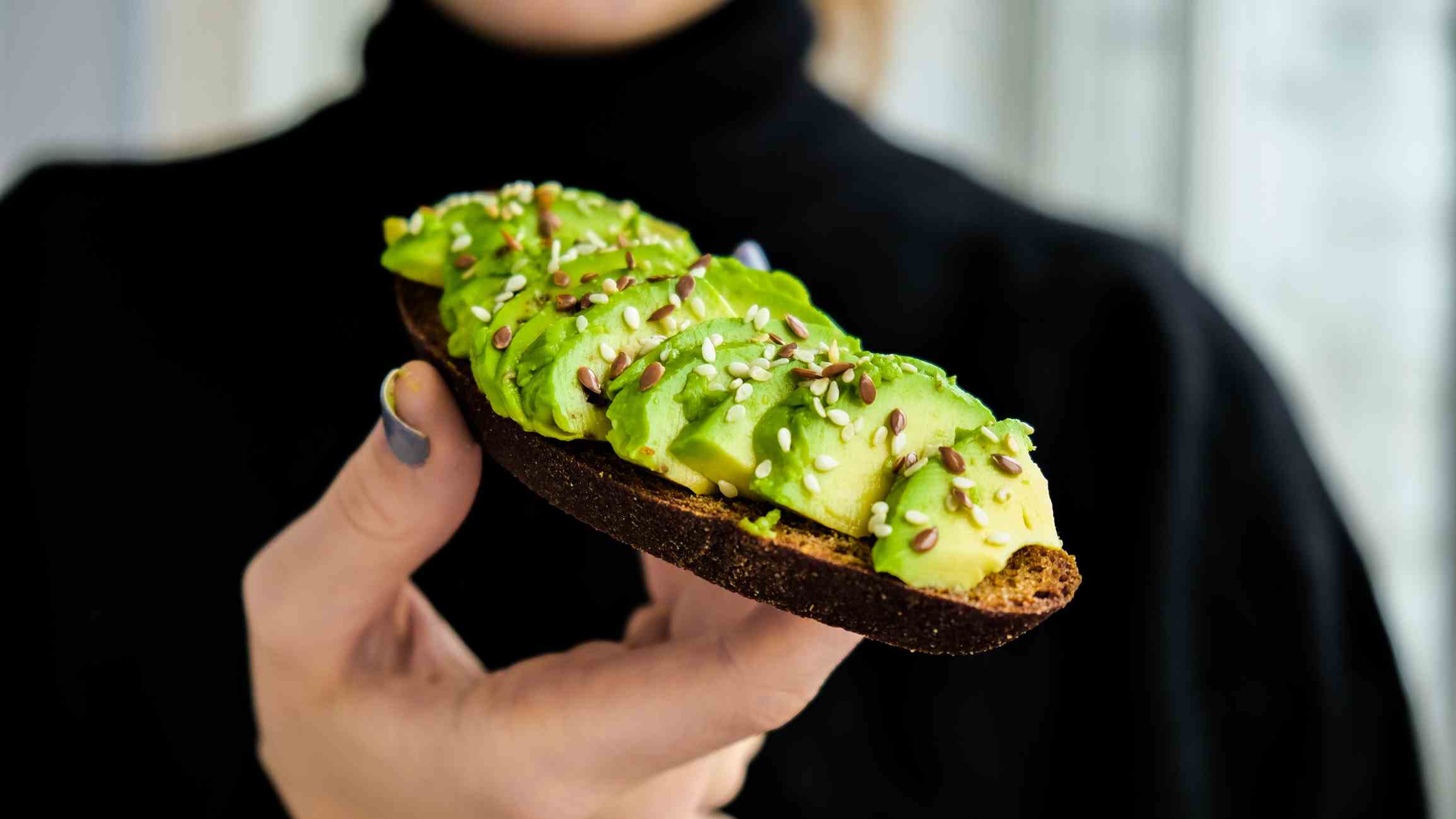 Hand, die Avocado-Toast hält.  Reife Hass-Avocado, Vollkornbrot, Sesam-Leinsamen.