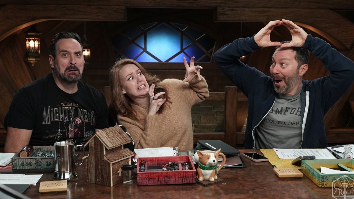 Zwei Männer und eine Frau schneiden an einem Tisch Gesichter in einem Werbefoto aus Critical Role: Campaign 3, Folge 83.