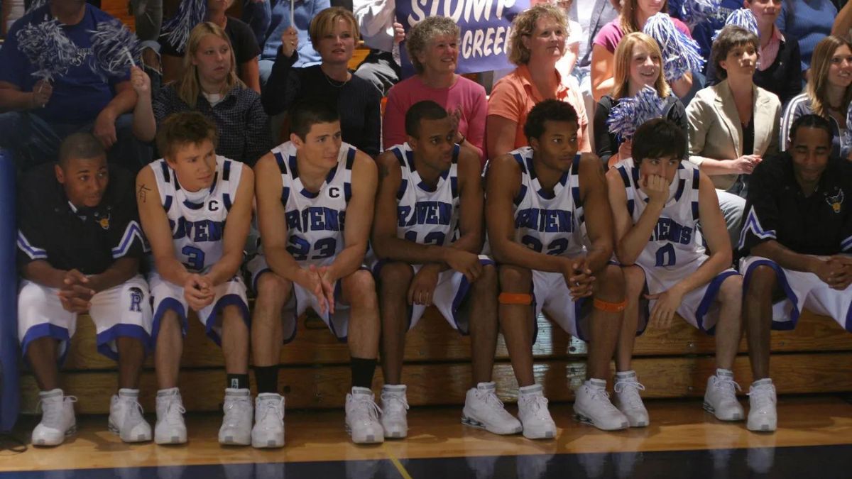 Mehrere Basketballspieler sitzen auf einer Bank neben einem Basketballplatz in One Tree Hill, hinter ihnen eine Menschenmenge